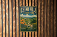 A vintage metal sign for Chinook Hills State Park, featuring a scenic landscape with hills, trees, and a winding road.