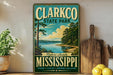 A vintage-style metal sign for Clarkco State Park, featuring a scenic landscape with a lake and trees, is displayed on a shelf next to a wooden cutting board and some lemons.