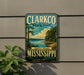 A vintage-style sign for Clarkco State Park, featuring a scenic landscape with a lake and trees, mounted on a wall next to a plant.