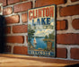 A vintage-style metal sign for Clinton Lake, Illinois, featuring a scenic image of a lake with a sailboat and mountains in the background.