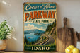 A vintage-style metal sign for Coeur d'Alene Parkway, featuring a scenic mountain landscape with a winding road and a boat on the water.