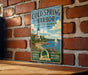 A vintage metal sign for Cold Spring Harbor State Park in New York, featuring a lighthouse and a scenic landscape with a sailboat on the water.