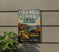 A vintage-style metal sign for Columbia Plateau Trail, located in Washington State Park, with a scenic mountain landscape in the background.