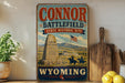a vintage-style sign for the "Connor Battlefield State Historic Site" in Wyoming, with a cowboy on horseback and a monument in the background.