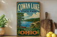 a vintage-style sign for Cowan Lake State Park, with a scenic landscape depicting a lake, trees, and mountains.