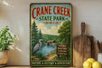 A vintage-style sign for Crane Creek State Park in Ohio, featuring a scenic illustration of a lake, trees, and a bird, along with text promoting outdoor activities such as hiking, boating, fishing, and camping.