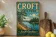 A vintage-style metal sign for Croft State Park, featuring a scenic landscape with a river, trees, and a lake, is displayed on a shelf next to a wooden cutting board and some potted plants.