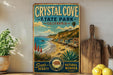 A vintage-style metal sign for Crystal Cove State Park, California, featuring a scenic beach landscape with palm trees, cacti, and a wooden pier.