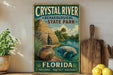 A vintage-style sign for Crystal River State Park in Florida, featuring an illustration of a manatee and a thatched-roof hut, with the text "Archaeological State Park" and "FLORIDA PRESERVE • PROTECT • DISCOVER" displayed prominently.