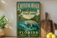 A vintage-style metal sign for Crystal River Preserve, Florida, featuring an illustration of a manatee in a body of water surrounded by palm trees and other natural scenery.