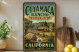 a vintage-style sign for Cuymacacho Ranch State Park in California, with a scenic illustration of a house, trees, and mountains. The sign is displayed on a wooden shelf, surrounded by various kitchen utensils and a potted plant.