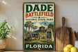 A vintage-style metal sign for Dade Battlefield Historic State Park in Florida, featuring a cannon and palm trees, is displayed on a shelf next to a wooden cutting board and a potted plant.