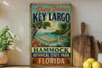 A vintage-style metal sign for "Key Largo" in the "Hammond Botanical State Park, Florida", featuring a hammock and tropical foliage.