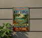 A vintage-style metal sign for Key Largo, Florida, featuring a hammock and tropical foliage, mounted on a wall.