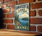 A vintage metal sign for Damariscotta Lake State Park in Maine, featuring a scenic landscape with a lake, mountains, and pine trees.