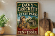 A vintage-style sign for Davy Crockett Birthplace State Park in Lawrenceburg, Tennessee, featuring an illustration of a man holding a rifle and standing in front of a cabin.