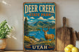 A vintage-style sign for Deer Creek State Park in Utah, featuring a scenic landscape with mountains, a lake, and a deer.