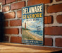 A vintage metal sign for Delaware Shore State Park, featuring a lighthouse and beach scene, is displayed on a brick wall.