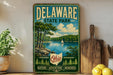 A vintage-style metal sign for Delaware State Park, featuring a scenic lake and mountains with a sailboat, is displayed on a wooden shelf next to a potted plant and a cutting board.