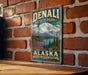 A vintage metal sign for Denali State Park in Alaska, featuring a mountain landscape and pine trees.