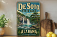 A vintage-style sign for DeSoto State Park in Alabama, featuring a scenic waterfall and forest landscape.