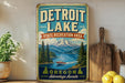 A vintage-style sign for Detroit Lake, a state recreation area in Oregon, featuring a scenic mountain landscape with a lake and boat.