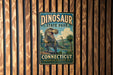 A vintage-style sign for Dinosaur State Park in Connecticut, featuring a T-Rex dinosaur and the text "DINOSAUR STATE PARK CONNECTICUT DISCOVER OUR PREHISTORIC PAST".
