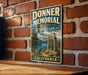 A vintage-style metal sign for Donner Memorial State Park in California, featuring a scenic landscape with a monument and mountains in the background.