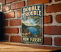 A vintage-style metal sign for Double Trouble State Park in New Jersey, featuring a scenic landscape with a lake and mountains.