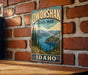 A vintage-style metal sign for Dworshak State Park in Idaho, featuring a scenic landscape with mountains, pine trees, and a lake.