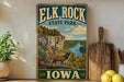 A vintage-style sign for Elk Rock State Park in Iowa, featuring a scenic landscape with a river, cliffs, and a deer.
