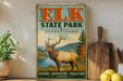 A vintage-style metal sign for Elk State Park in Pennsylvania, featuring an illustration of a deer in a scenic landscape.