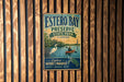 A vintage metal sign for Estero Bay Preserve State Park in Florida, featuring a scenic illustration of a person kayaking and a bird, mounted on a wooden wall.