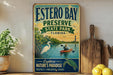 A vintage-style metal sign for Estero Bay Preserve, Florida, featuring an illustration of a person kayaking and a bird, along with the text "Explore Nature's Paradise" and "Protect - Preserve - Enjoy".