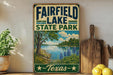 a vintage-style sign for Fairfield Lake State Park in Texas, with a scenic landscape depicting a lake surrounded by trees and flowers.