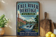 a vintage-style sign for the Fall River Heritage State Park in Massachusetts, with a scenic illustration of a river, bridge, and waterfall. The sign is displayed on a shelf next to a wooden cutting board and a potted plant, creating a rustic and charming atmosphere.
