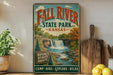 a vintage-style sign for Fall River State Park in Kansas, with a waterfall and forest scenery. The sign is displayed on a shelf next to a wooden cutting board and a potted plant.
