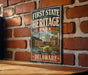 A vintage-style metal sign for First State Heritage Park in Delaware, featuring an illustration of a historic building and ship, mounted on a brick wall.