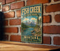 A vintage-style metal sign for Fish Creek State Park in Montana, featuring a scenic mountain landscape with a river and pine trees.