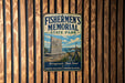 A vintage metal sign for Fishermen's Memorial State Park, featuring an illustration of a lighthouse and a monument, mounted on a wooden wall.