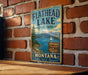 A vintage-style metal sign for Flathead Lake State Park, featuring a scenic landscape with mountains, a lake, and a sailboat.