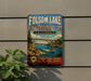 A vintage metal sign for Folsom Lake State Recreation Area in California, featuring a scenic illustration of a bridge over a river with boats and people enjoying the outdoors.