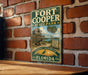 A vintage-style metal sign for Fort Cooper State Park in Florida, featuring a palm tree and a wooden fort, is displayed on a brick wall.
