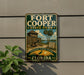 A vintage-style sign for Fort Cooper State Park, featuring a wooden building and palm trees, is displayed on a wall next to a plant.