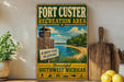 A vintage-style sign for the Fort Custer Recreation Area in Southwest Michigan, featuring a beach scene with a lighthouse and a wooden guardhouse.