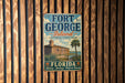 A vintage-style metal sign for Fort George Island, Florida, featuring a castle and palm trees, mounted on a wooden wall.
