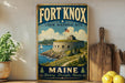 A vintage-style metal sign for the Fort Knox State Historic Site in Maine, featuring an illustration of a fort on a rocky coastline with a sailboat in the background.
