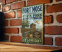 A vintage-style sign for Fort Mose Historic State Park in Florida, featuring a brick wall backdrop and a wooden table.