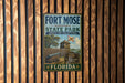 A vintage-style metal sign for Fort Mose Historic State Park in Florida, featuring a wooden fence and a cannon, mounted on a wooden wall.