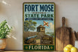 A vintage-style metal sign for Fort Mose Historic State Park in Florida, featuring an illustration of a cannon and a wooden fort.